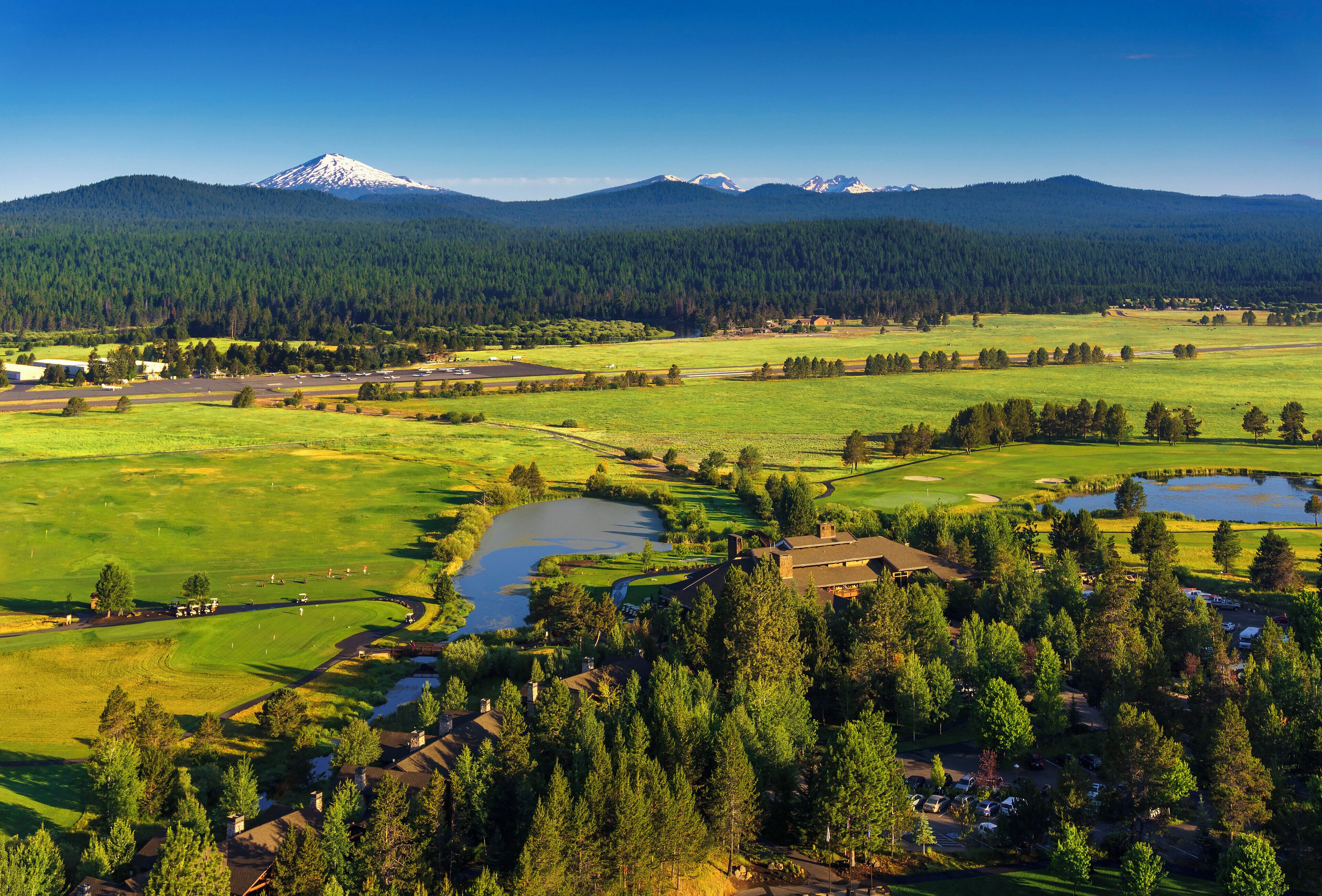 Un campo de golf con un lago, rodeado de bosques densos y montañas al fondo.