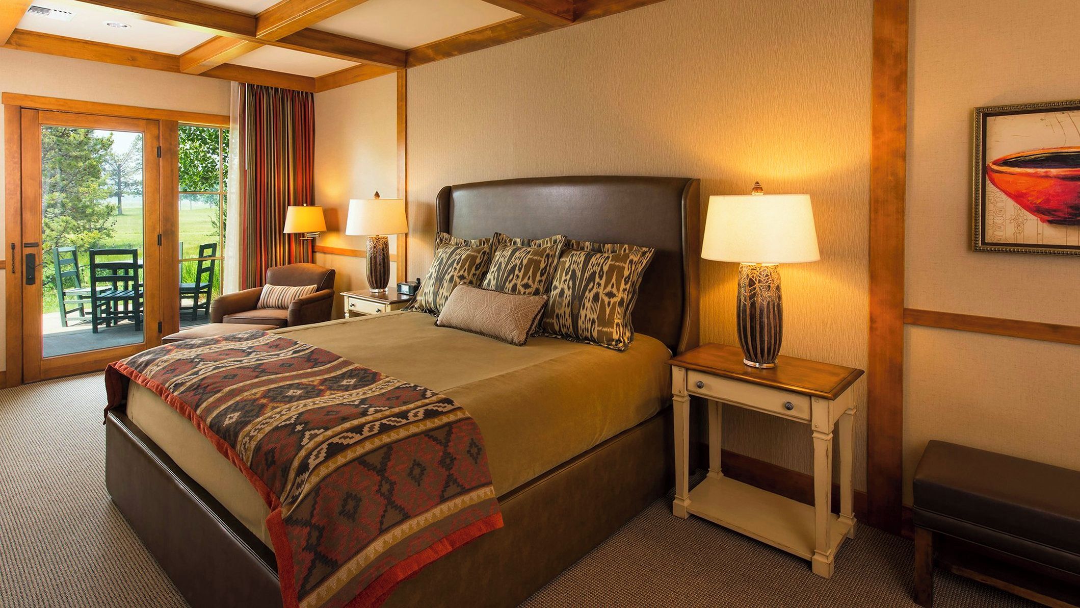 A hotel room with a large bed, a bedside table with a lamp, a chair, and a view of the outdoors through a sliding glass door.