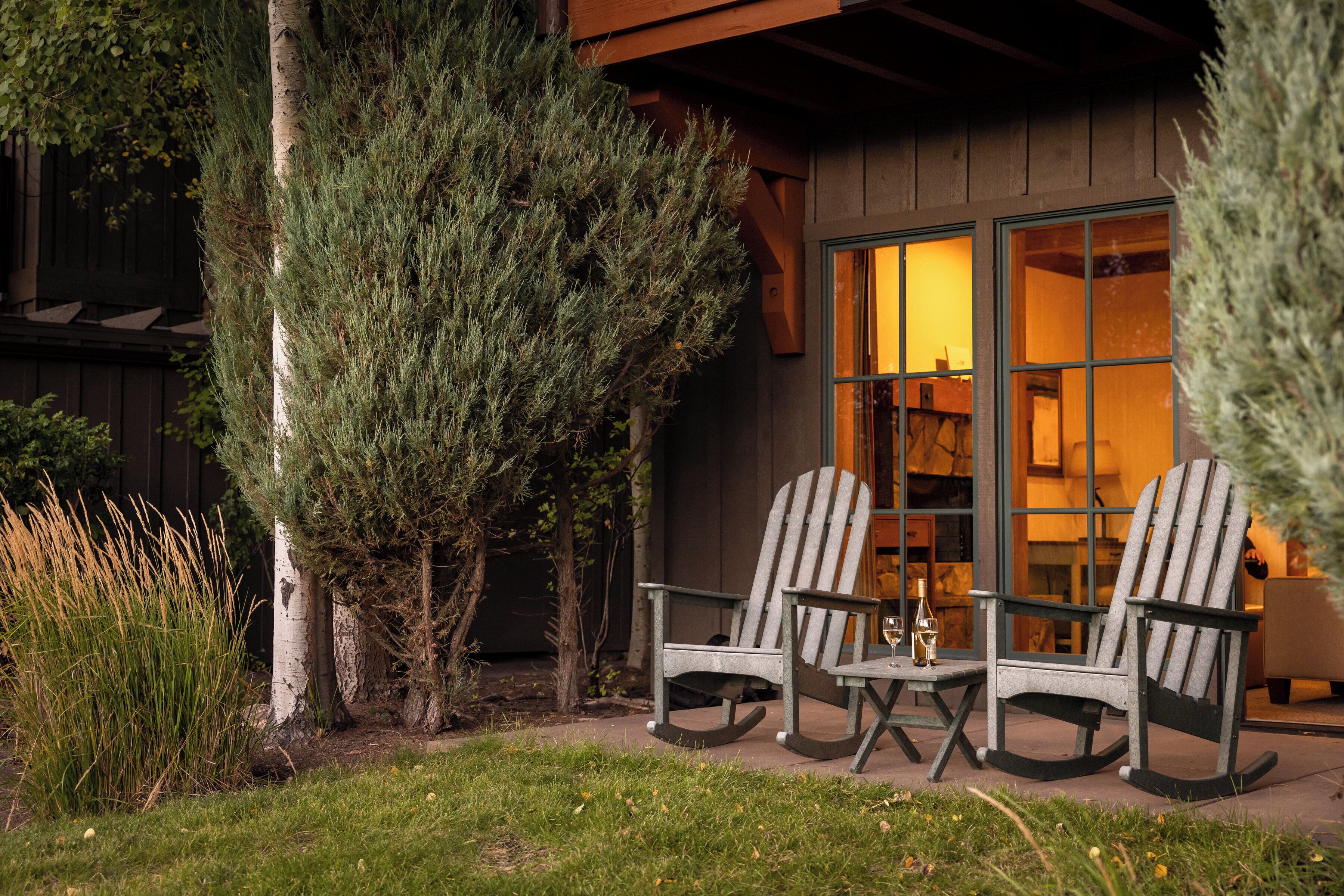 A wooden cabin with two rocking chairs and a table, surrounded by greenery.
