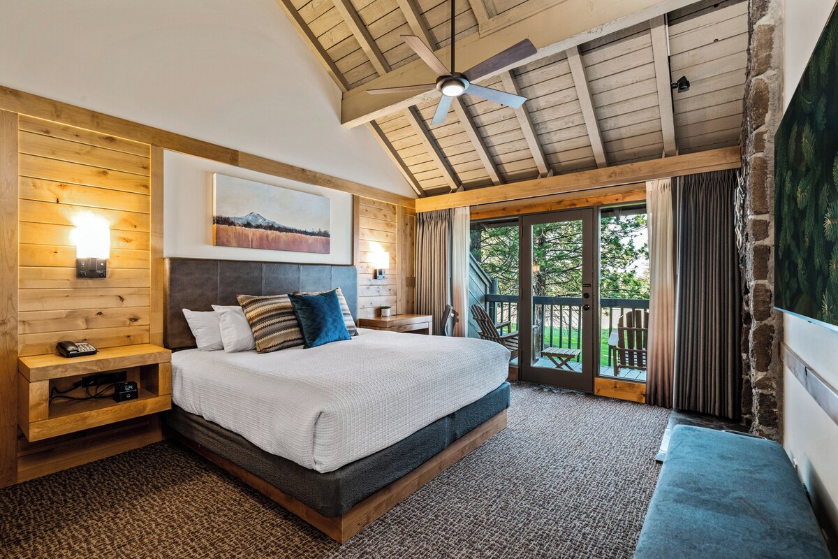 Une chambre avec un plafond en bois, un lit avec une literie blanche, une table de chevet avec une lampe, et une vue sur l’extérieur par une porte vitrée.