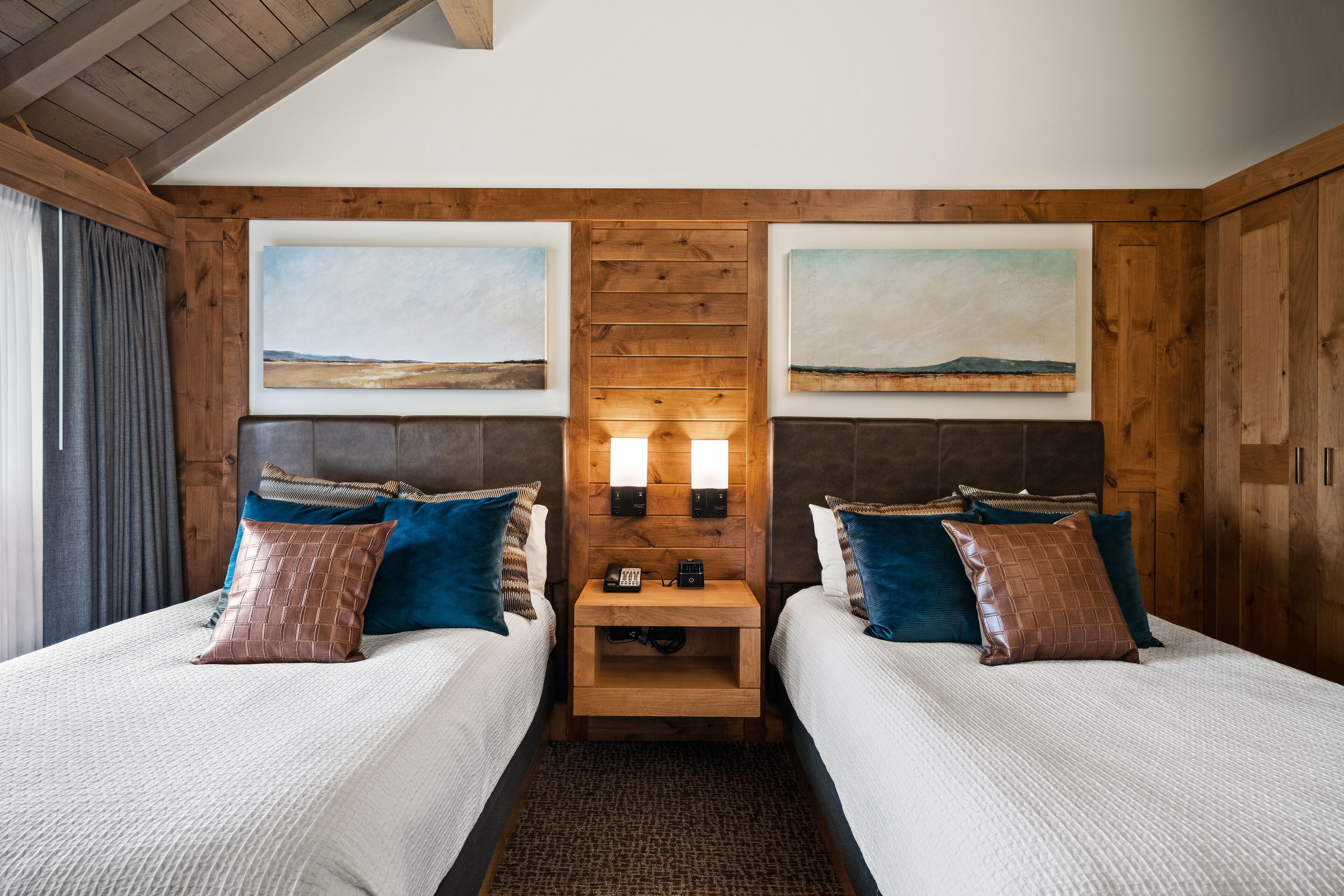 A hotel room with two beds, wooden headboards, a nightstand with a phone, and two wall-mounted lamps.