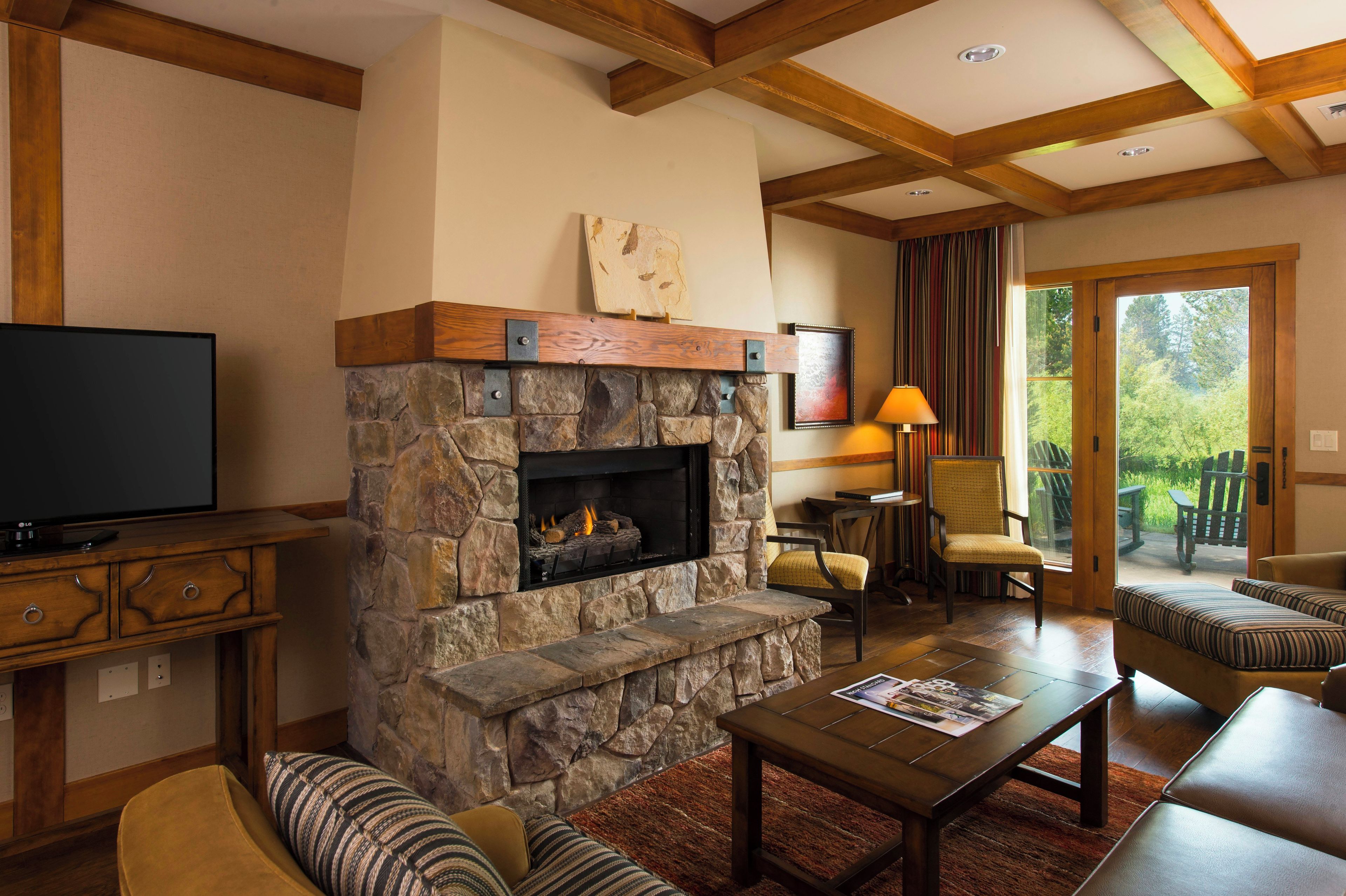 A living room with a stone fireplace, wooden beams, a television, and a view of the outdoors.