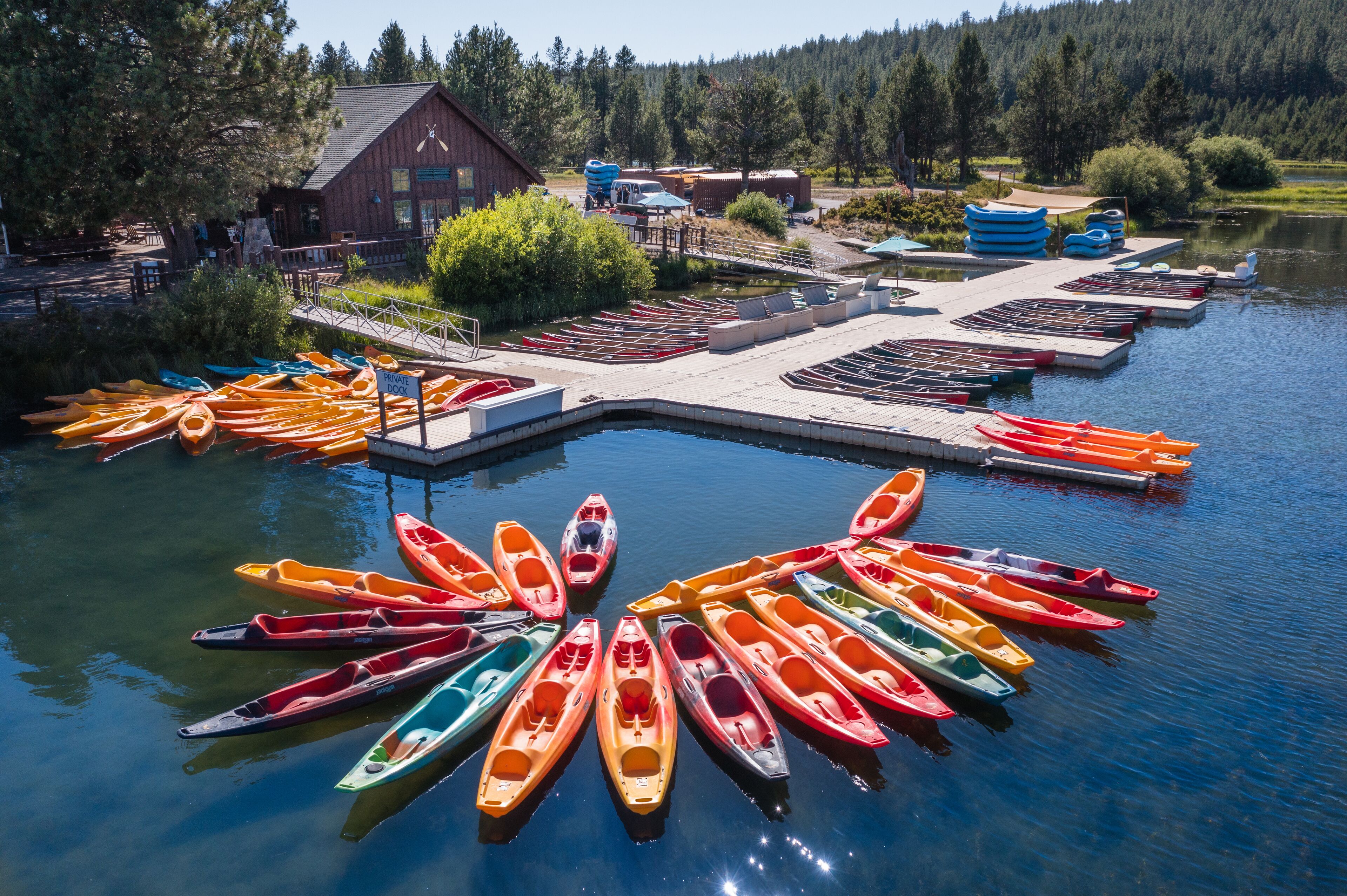 Un muelle con kayaks de colores dispuestos en semicírculo, un edificio de madera y un área boscosa al fondo.
