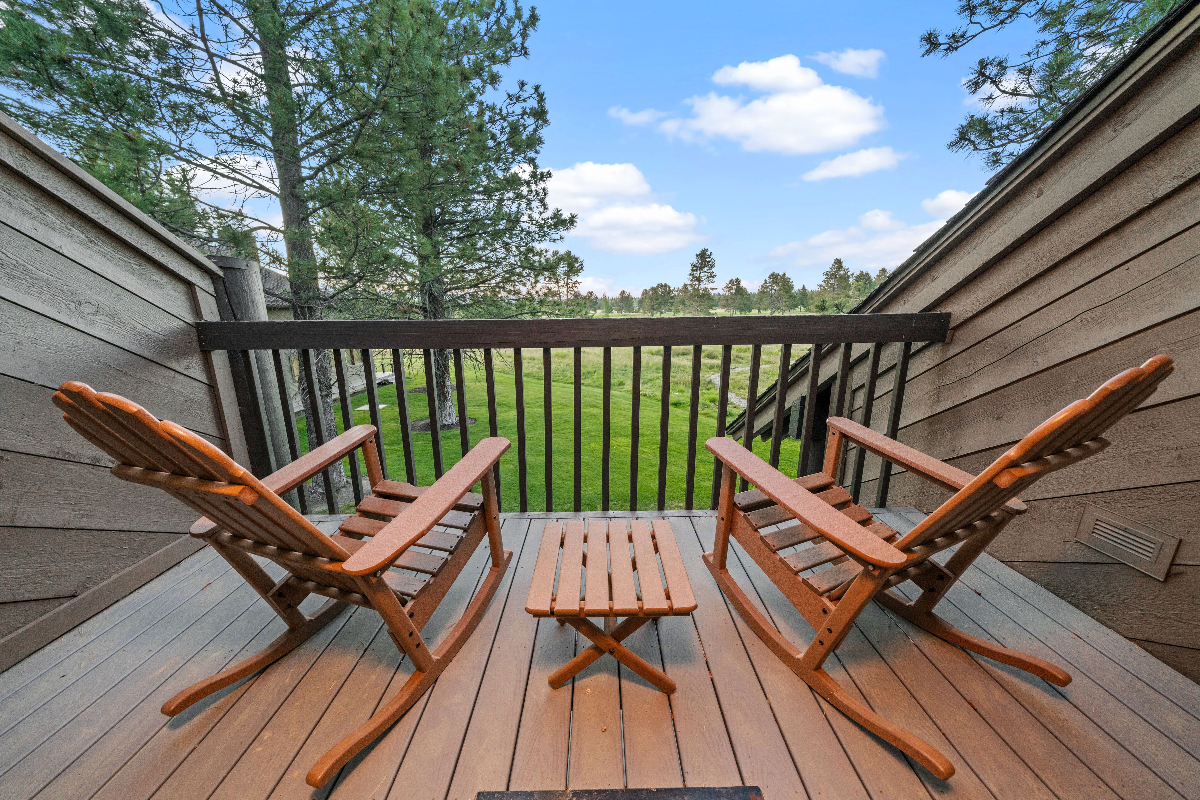 Une terrasse en bois avec deux fauteuils à bascule et une petite table, donnant sur une cour verdoyante bordée d’arbres.