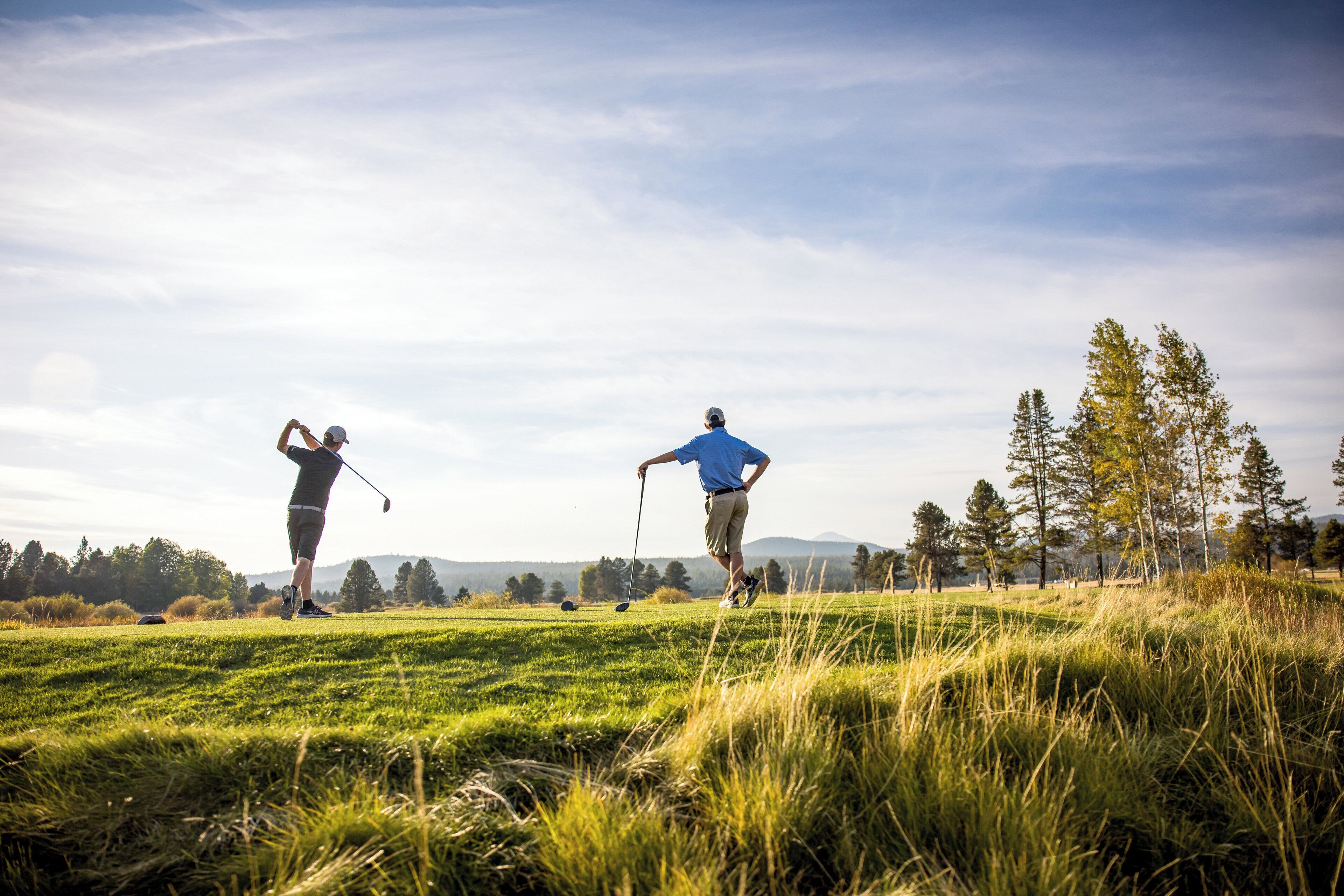 Deux personnes jouent au golf sur un terrain herbeux, avec des arbres et des collines en arrière-plan.