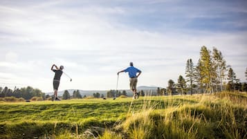 Deux personnes jouent au golf sur un terrain herbeux, avec des arbres et des collines en arrière-plan.