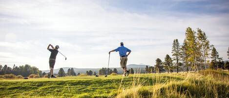 Två personer spelar golf på en gräsklädd plan med träd och kullar i bakgrunden.