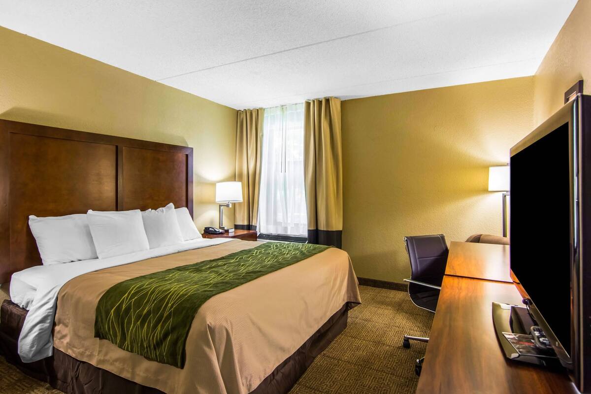 Une chambre d’hôtel avec un lit, un bureau avec un ordinateur, une télévision et une fenêtre avec des rideaux.