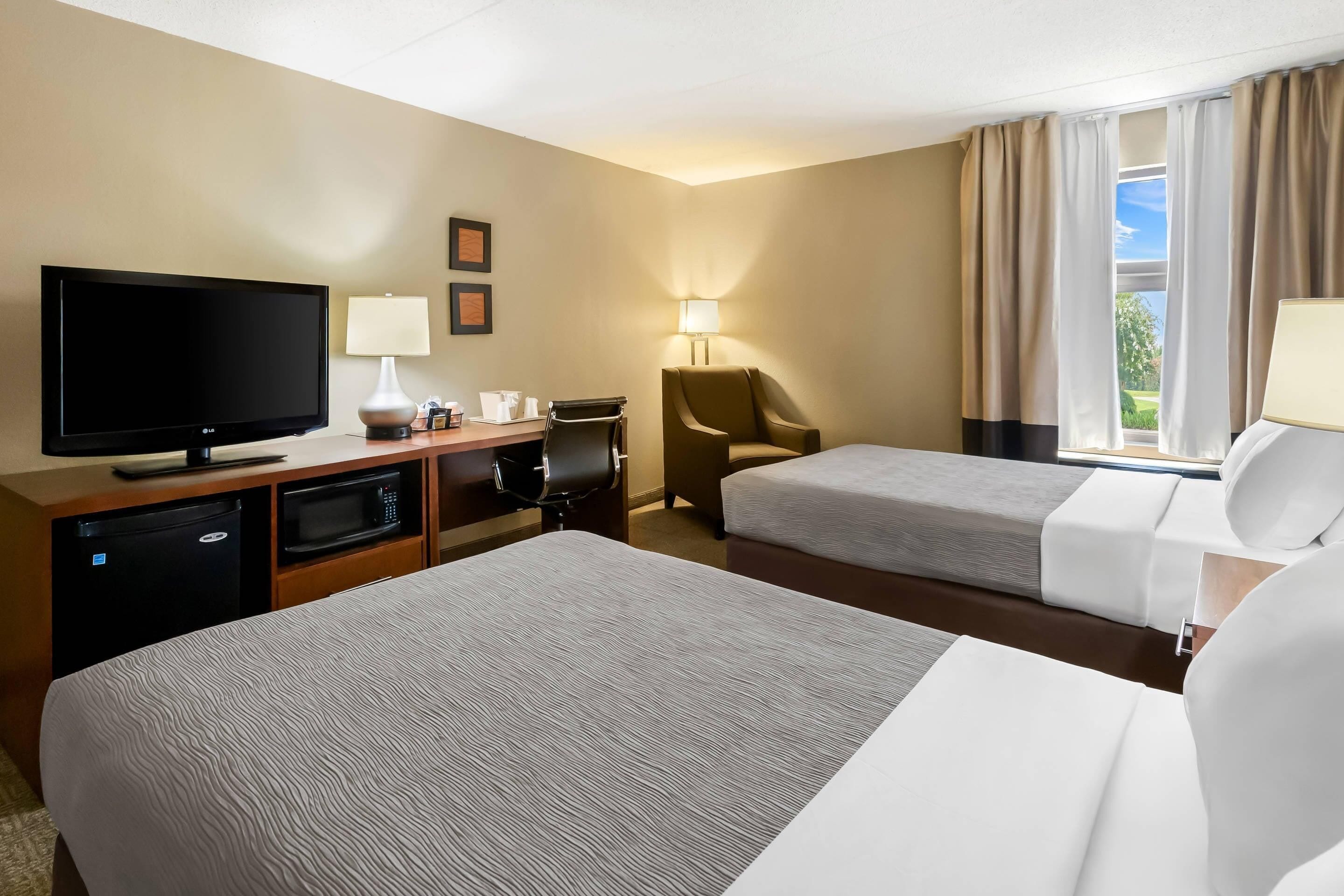 Habitación de hotel con dos camas, televisión, escritorio con computadora, una silla y ventana con cortinas.