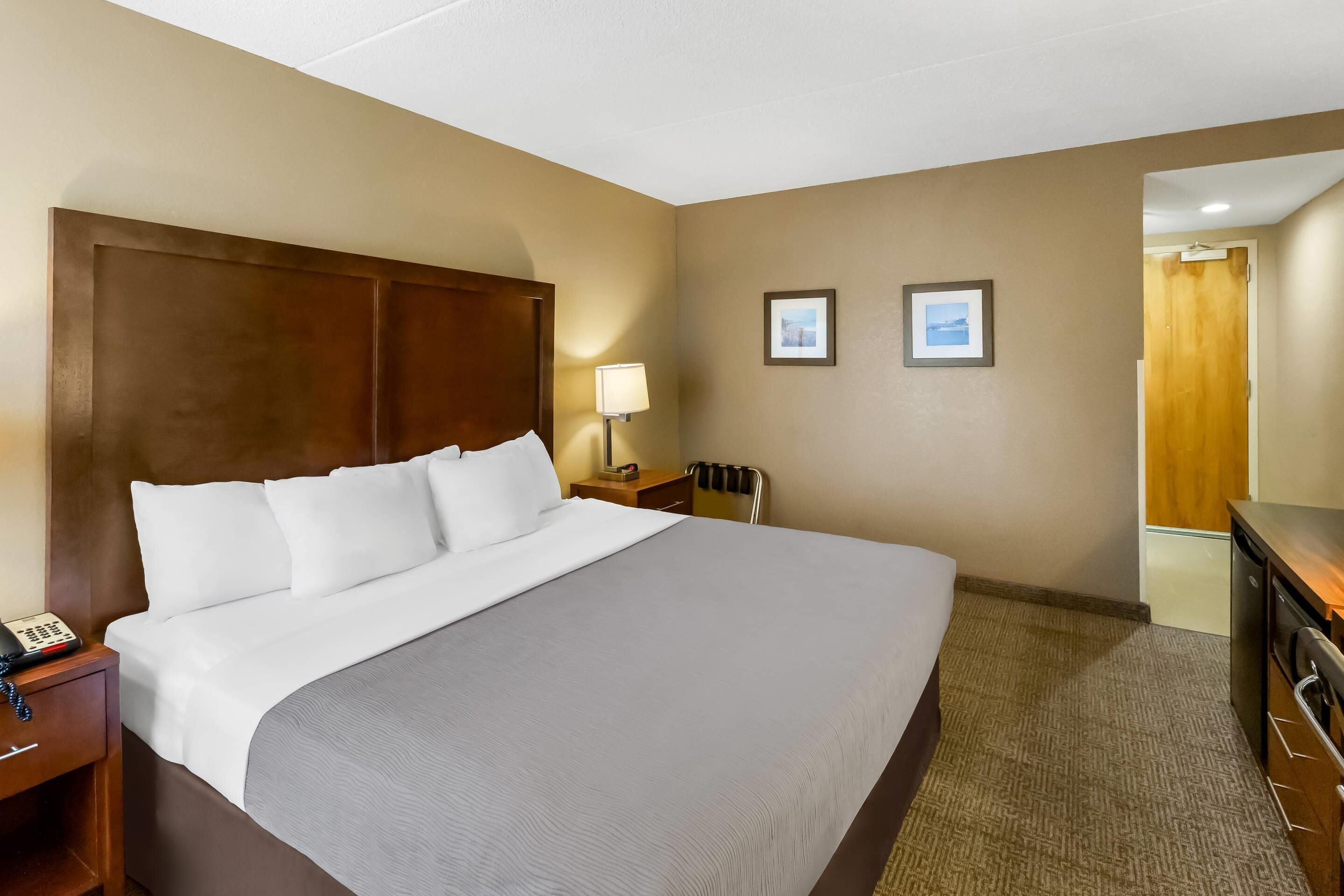 A hotel room with a large bed, a bedside table with a lamp, a chair, and a desk with a computer.