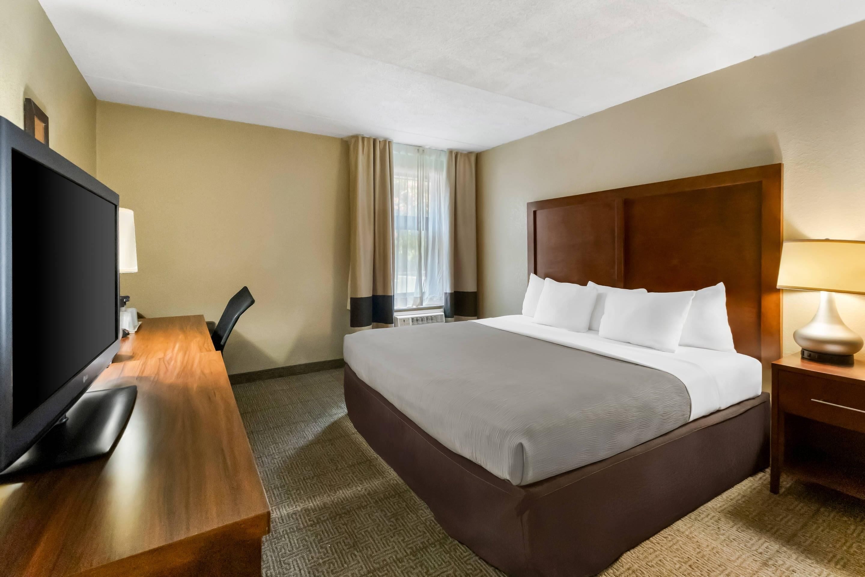 A hotel room with a bed, a desk with a television, a lamp, and a window with curtains.