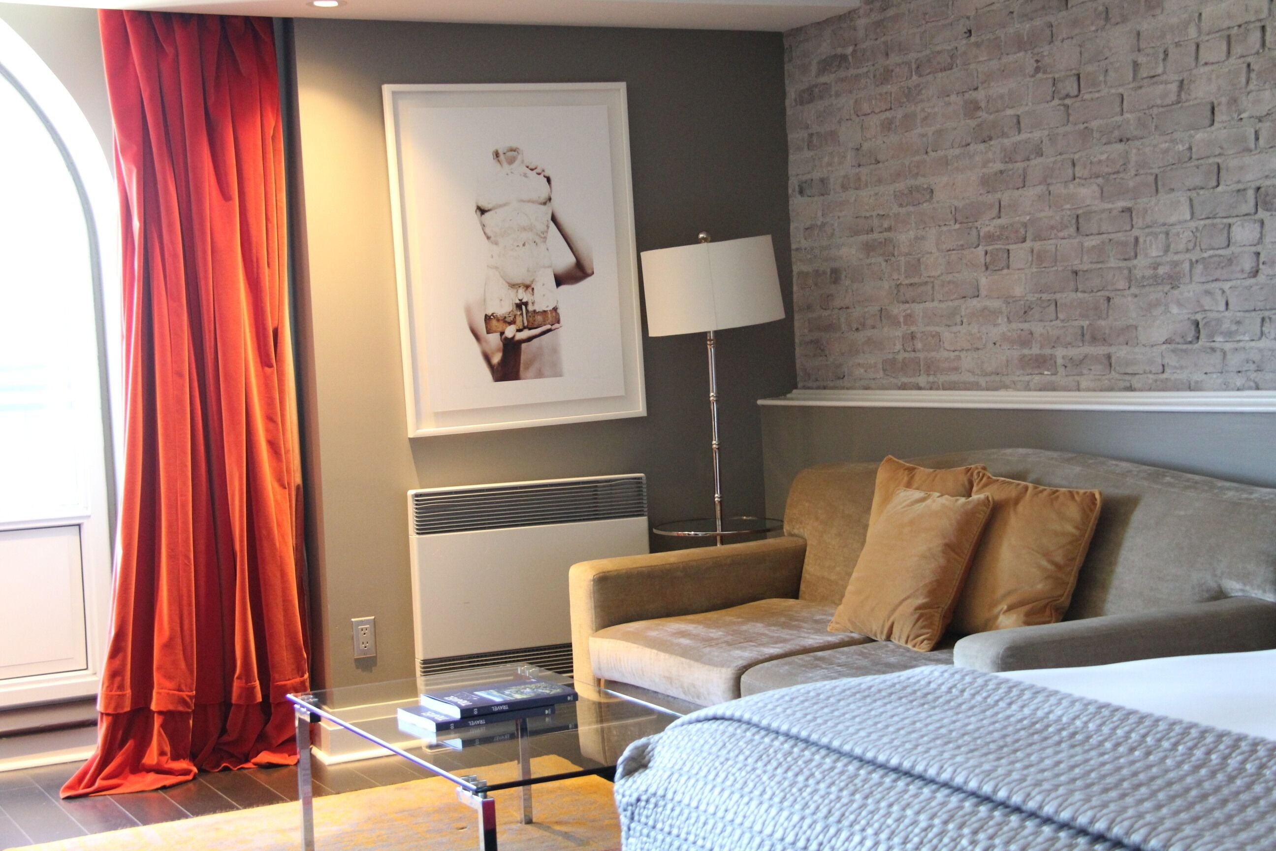 A modern hotel room with a grey brick wall, a beige sofa, a red curtain, a framed artwork, a standing lamp, and a glass coffee table.