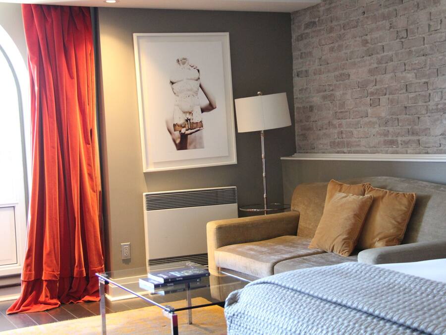 A modern hotel room with a grey brick wall, a beige sofa, a red curtain, a framed artwork, a standing lamp, and a glass coffee table.