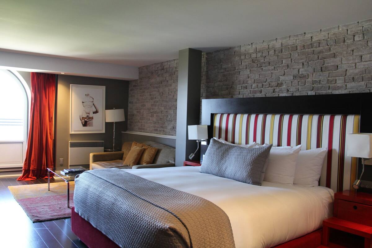 A modern hotel room with a large bed, a brick wall, a red accent wall, and a window with red curtains.