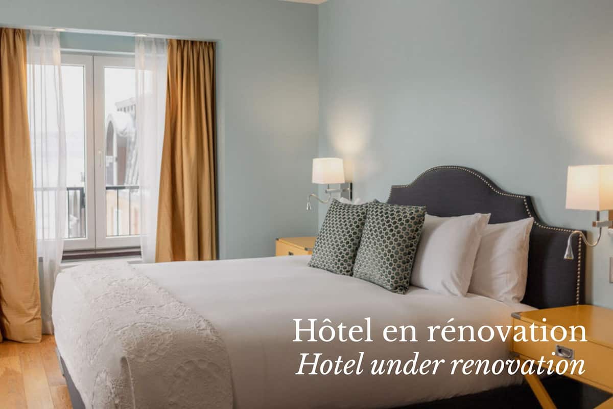 A hotel room under renovation with a large bed, two bedside tables with lamps, and a view through a window with curtains.