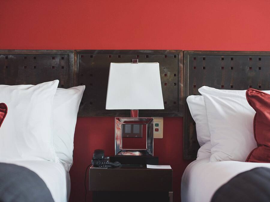 A hotel room with two beds, each with white bedding and red pillows, a nightstand with a lamp, and a red wall.