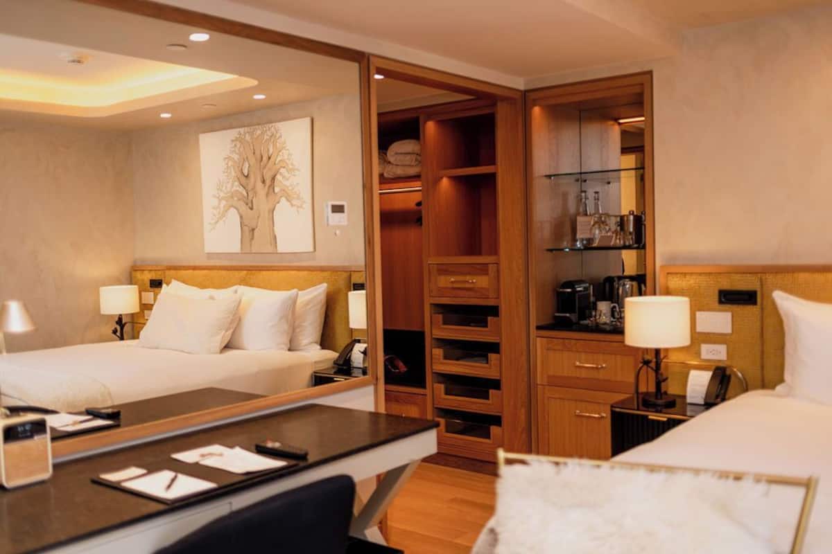 A hotel room with two beds, a desk, a mirror, a wardrobe, and a shelf with toiletries.