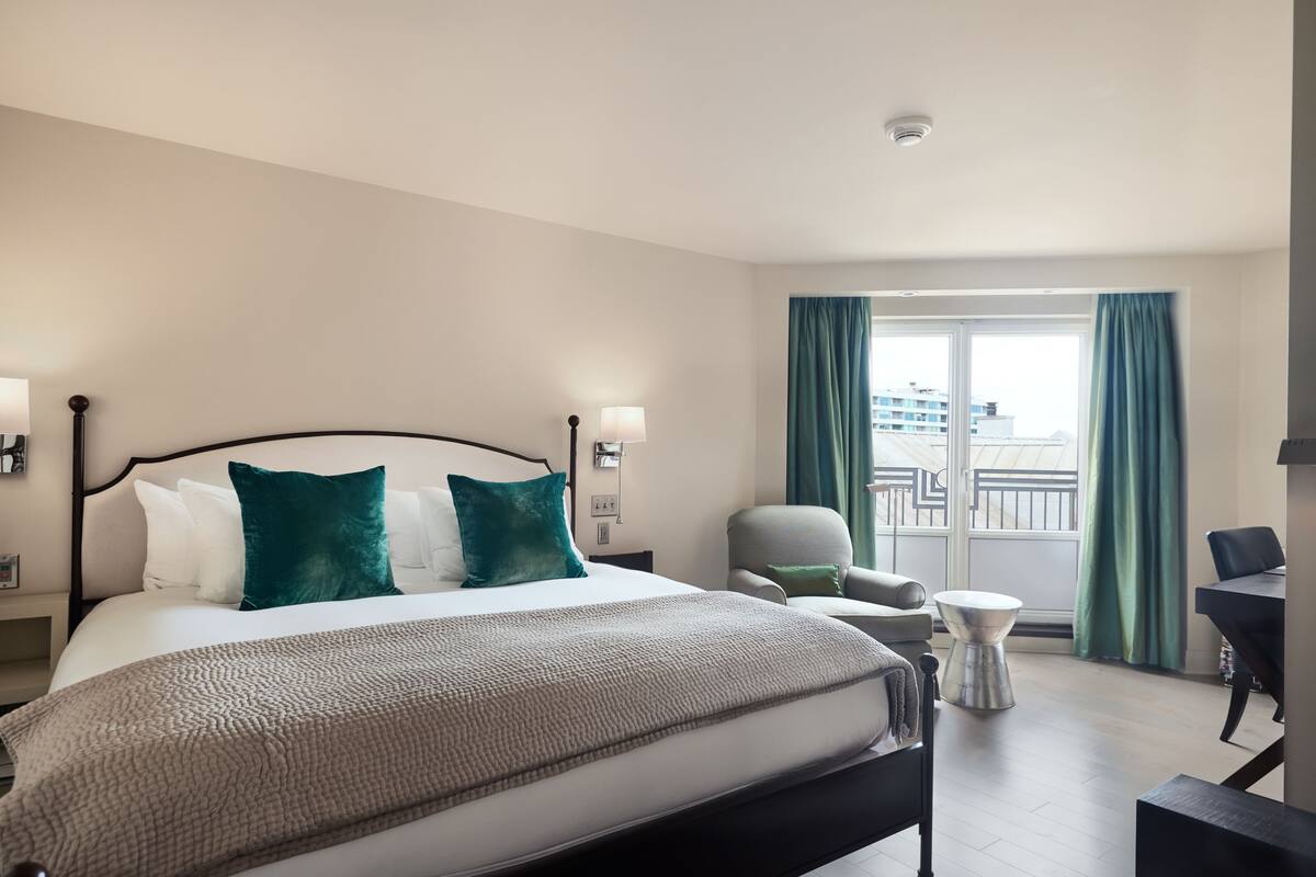 A hotel room with a large bed, two bedside tables, a chair, a small round table, and a view of a building through a window with curtains.