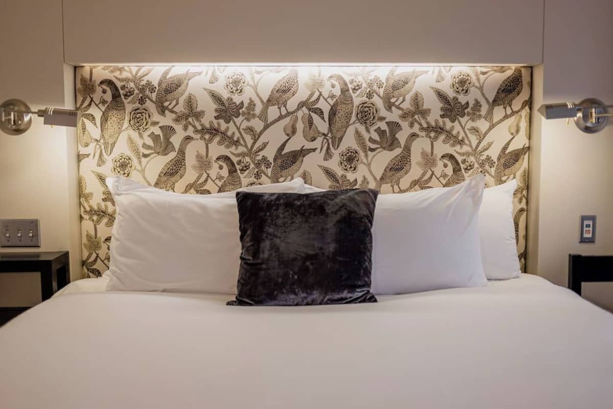 A neatly made bed with white linens and a black pillow, set against a wall with a floral wallpaper pattern.