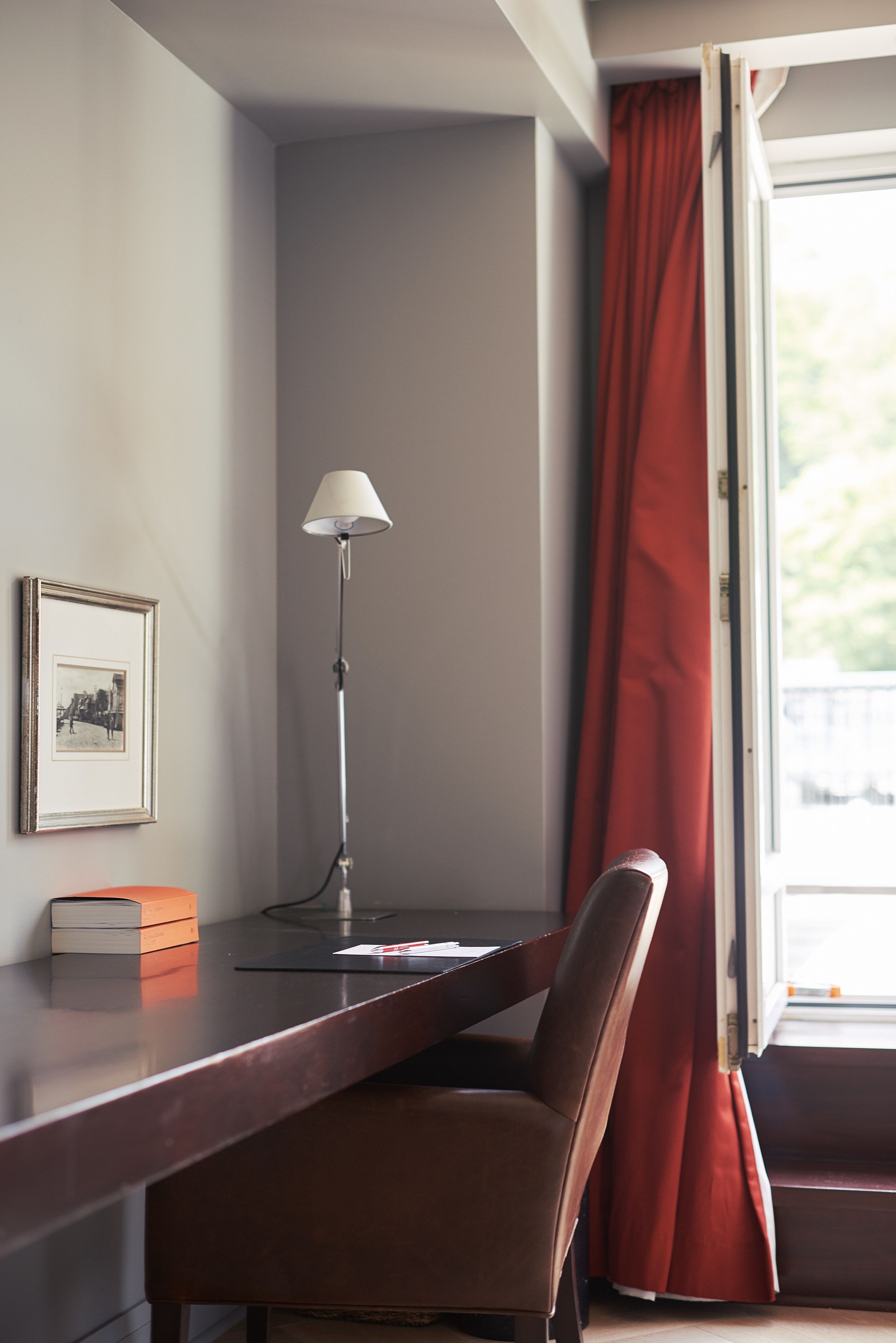 Una habitación con escritorio, silla, lámpara, cuadro enmarcado y ventana con cortinas rojas.