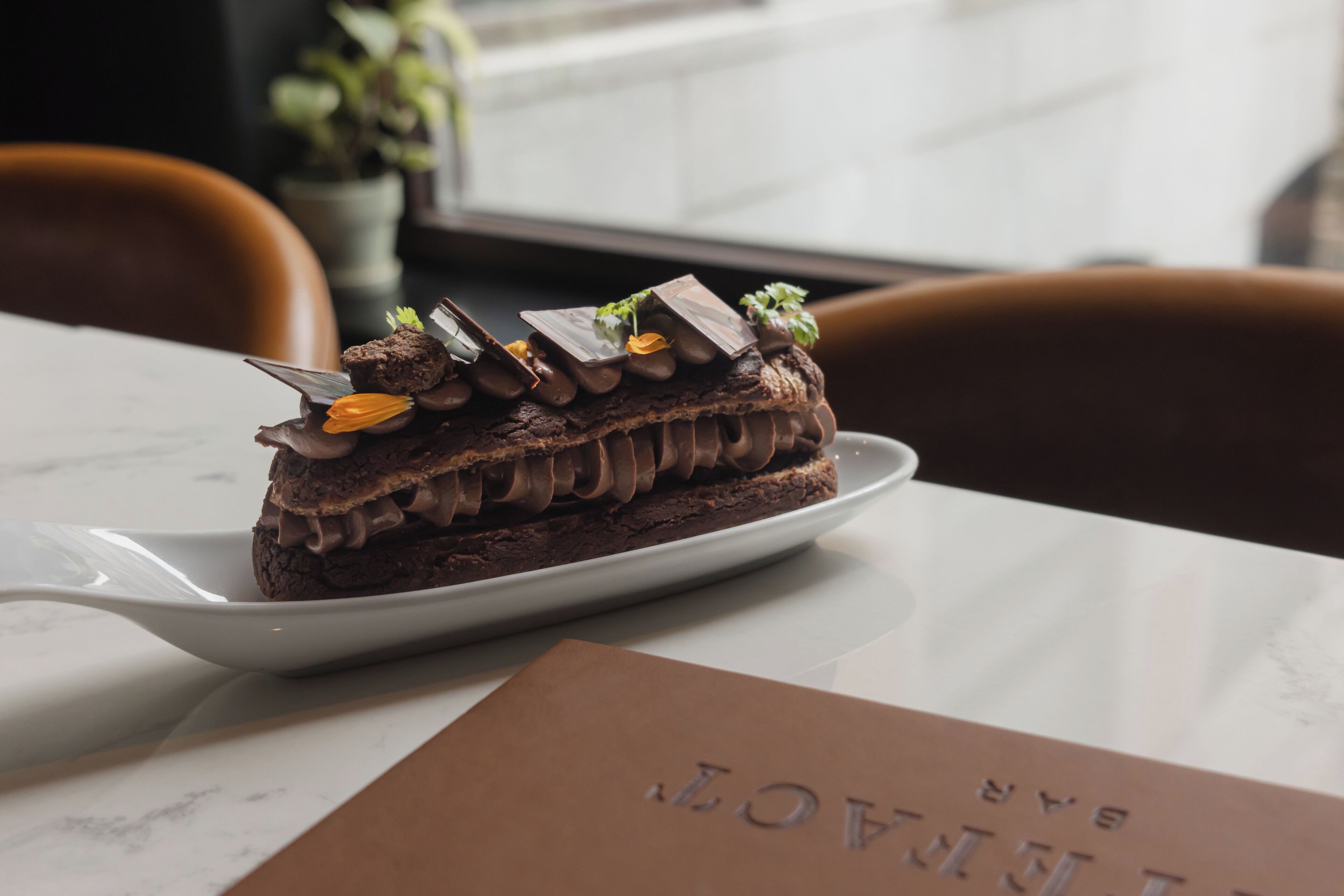 Una porción de pastel de chocolate sobre un plato blanco, decorado con trozos de chocolate y hojas encima, sobre una mesa de mármol.