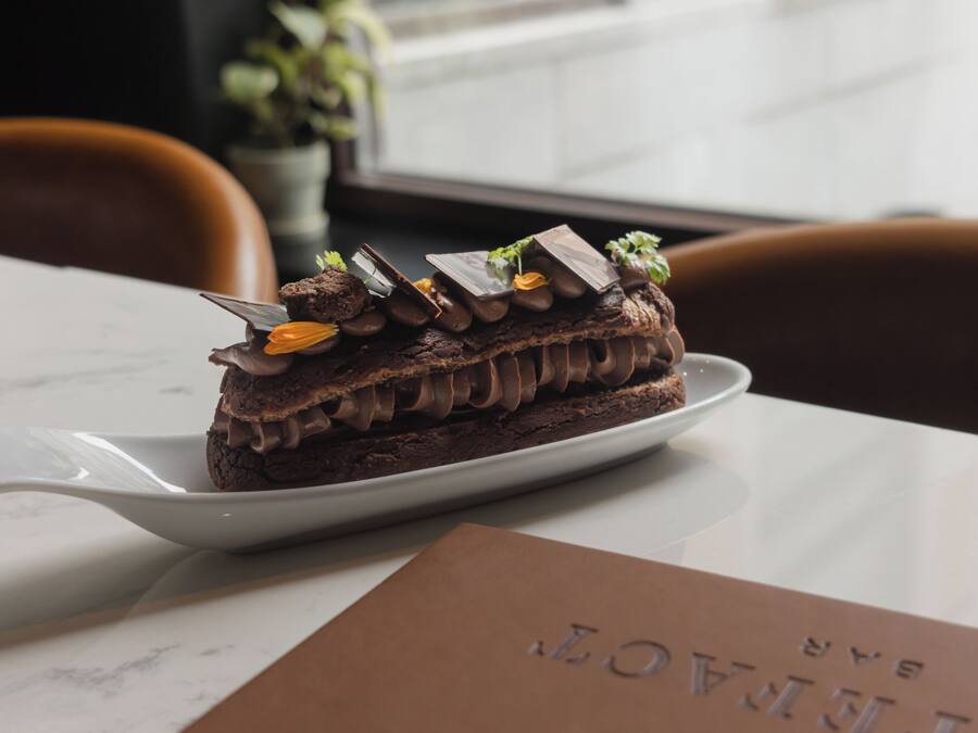 Una porción de pastel de chocolate sobre un plato blanco, decorado con trozos de chocolate y hojas encima, sobre una mesa de mármol.