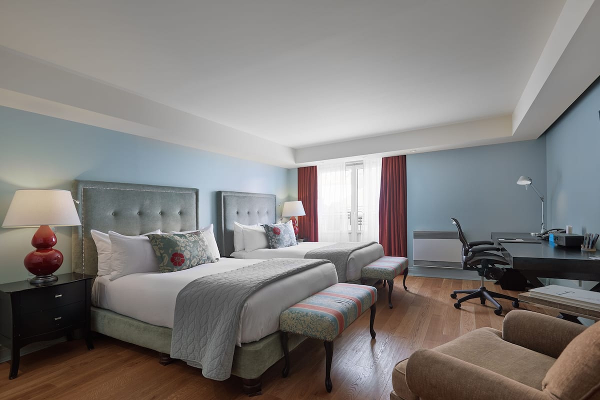 A hotel room with two beds, a desk, a chair, and a lamp.