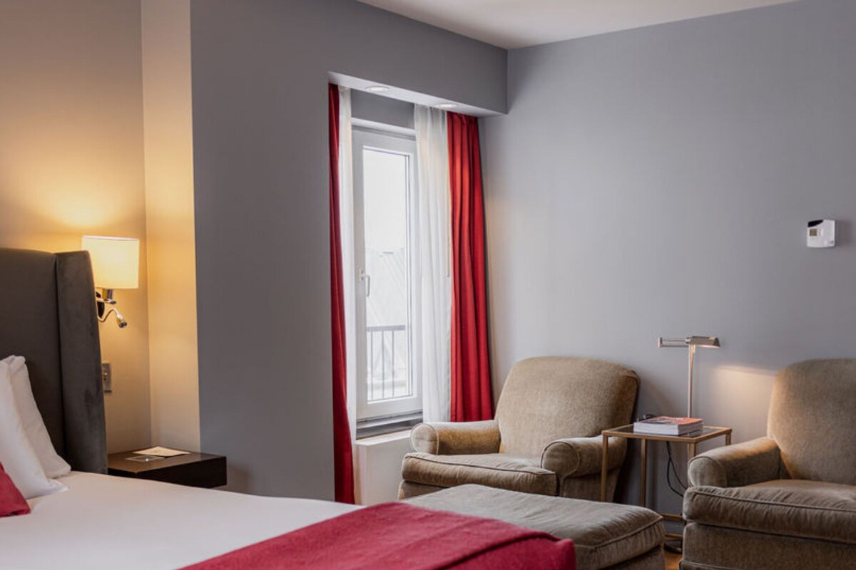 A hotel room under renovation with a bed, chairs, a small table, and a window with red curtains.