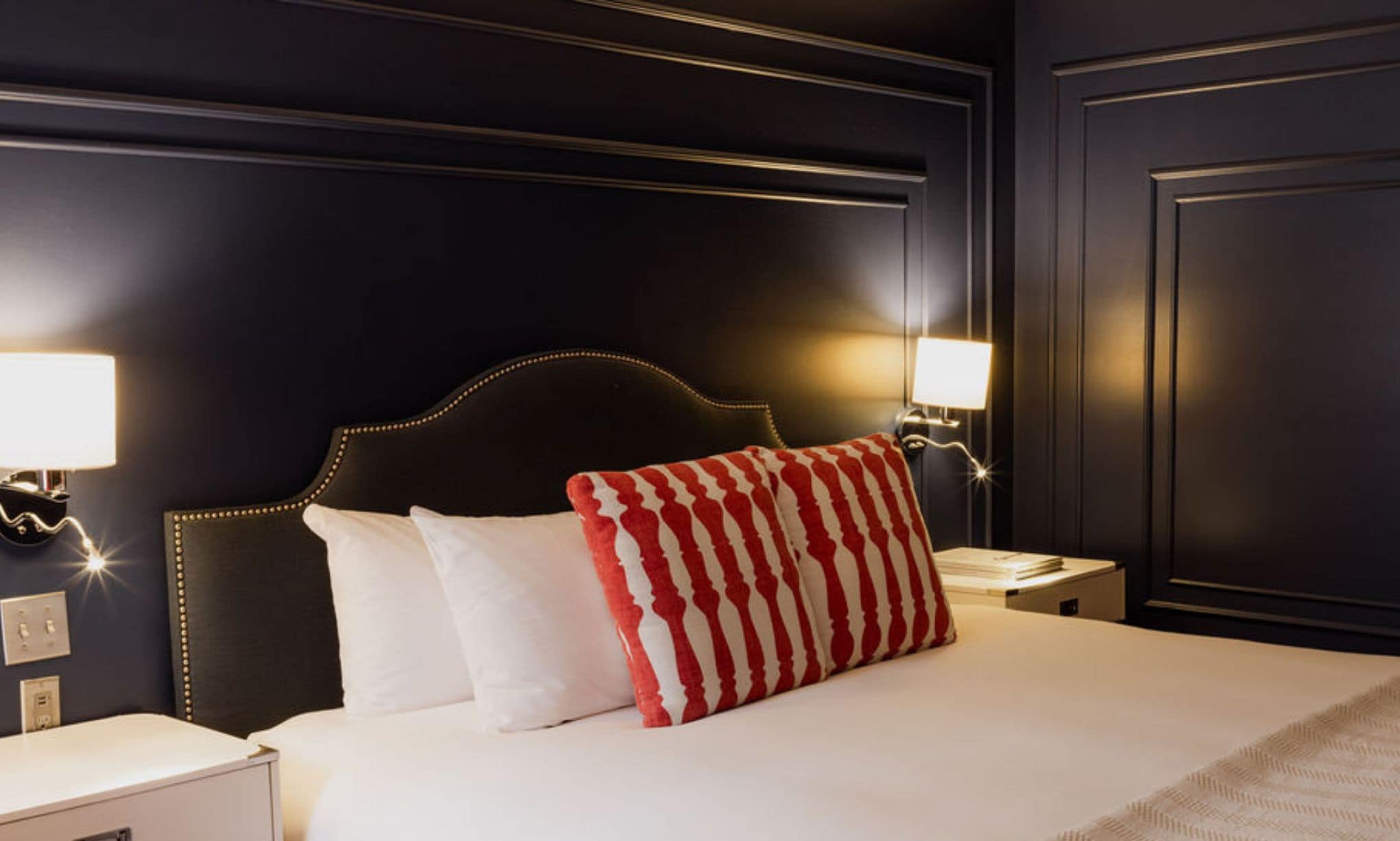 Quarto de hotel em reforma, com parede escura, cama com almofadas listradas em branco e vermelho, e abajures laterais.
