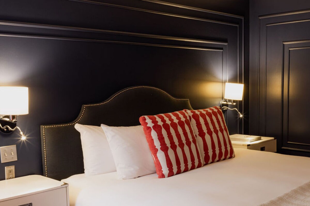 A hotel room under renovation with a dark wall, a bed with white and red striped pillows, and bedside lamps.
