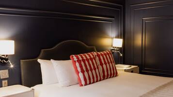 Quarto de hotel em reforma, com parede escura, cama com almofadas listradas em branco e vermelho, e abajures laterais.