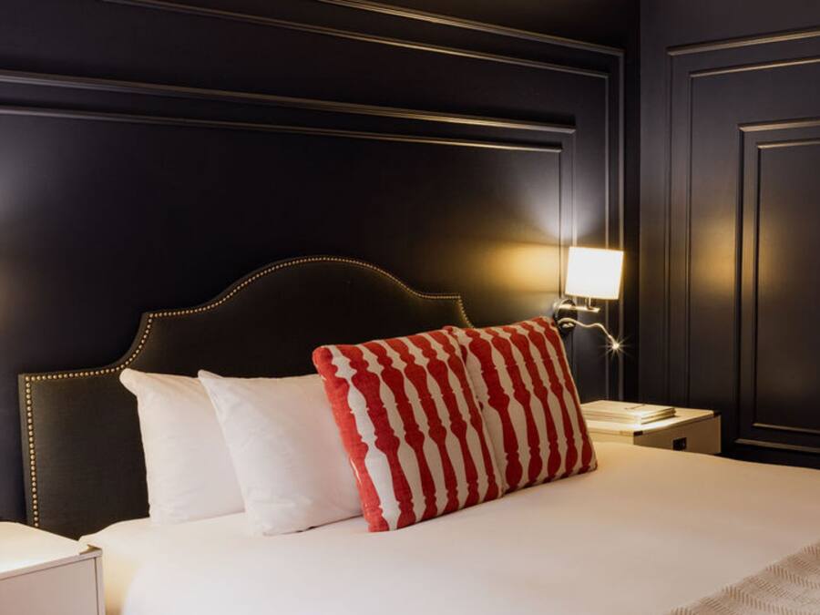 Quarto de hotel em reforma, com parede escura, cama com almofadas listradas em branco e vermelho, e abajures laterais.