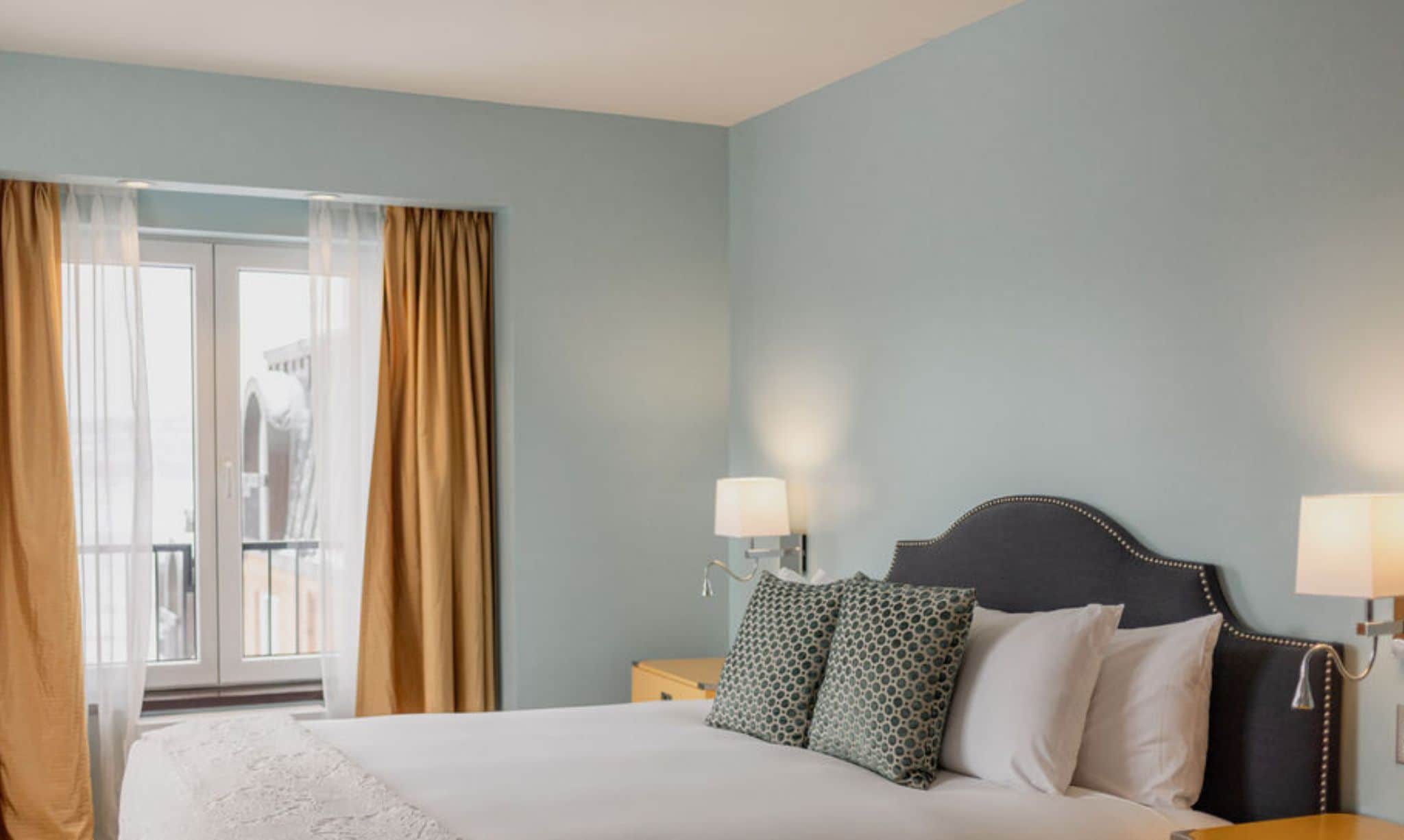 Habitación de hotel en renovación con una cama grande, dos mesitas de noche con lámparas y vistas a través de una ventana con cortinas.