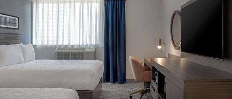 Habitación de hotel con dos camas, un escritorio, un televisor y un ventanal con cortinas.