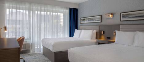 A hotel room with two beds, a desk, a chair, and large windows with sheer curtains.