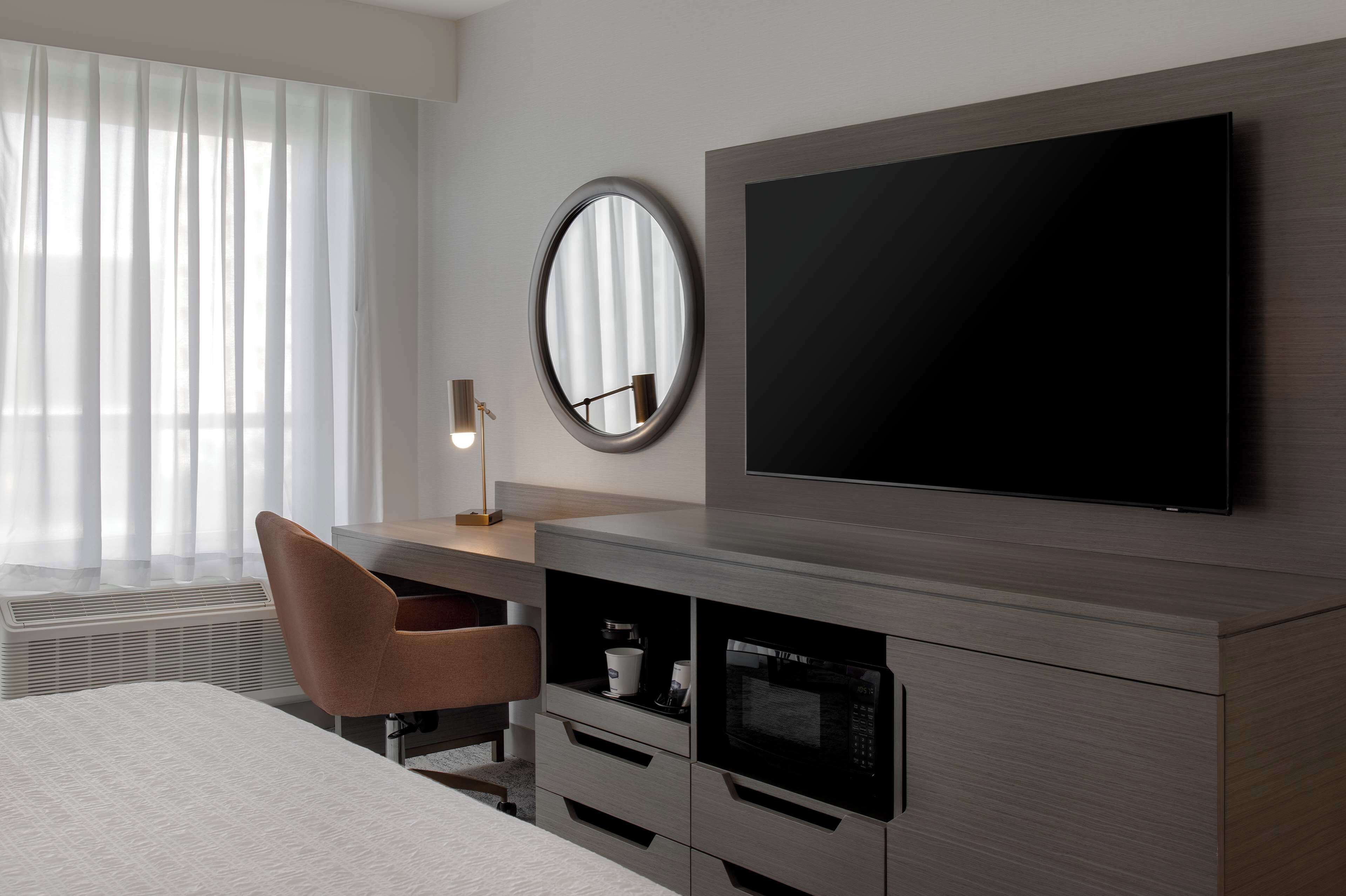 Een moderne hotelkamer met een grote flatscreen televisie aan de muur, een ronde spiegel, een bureau met lamp, een stoel en een ingebouwde magnetron en koffiezetapparaat.