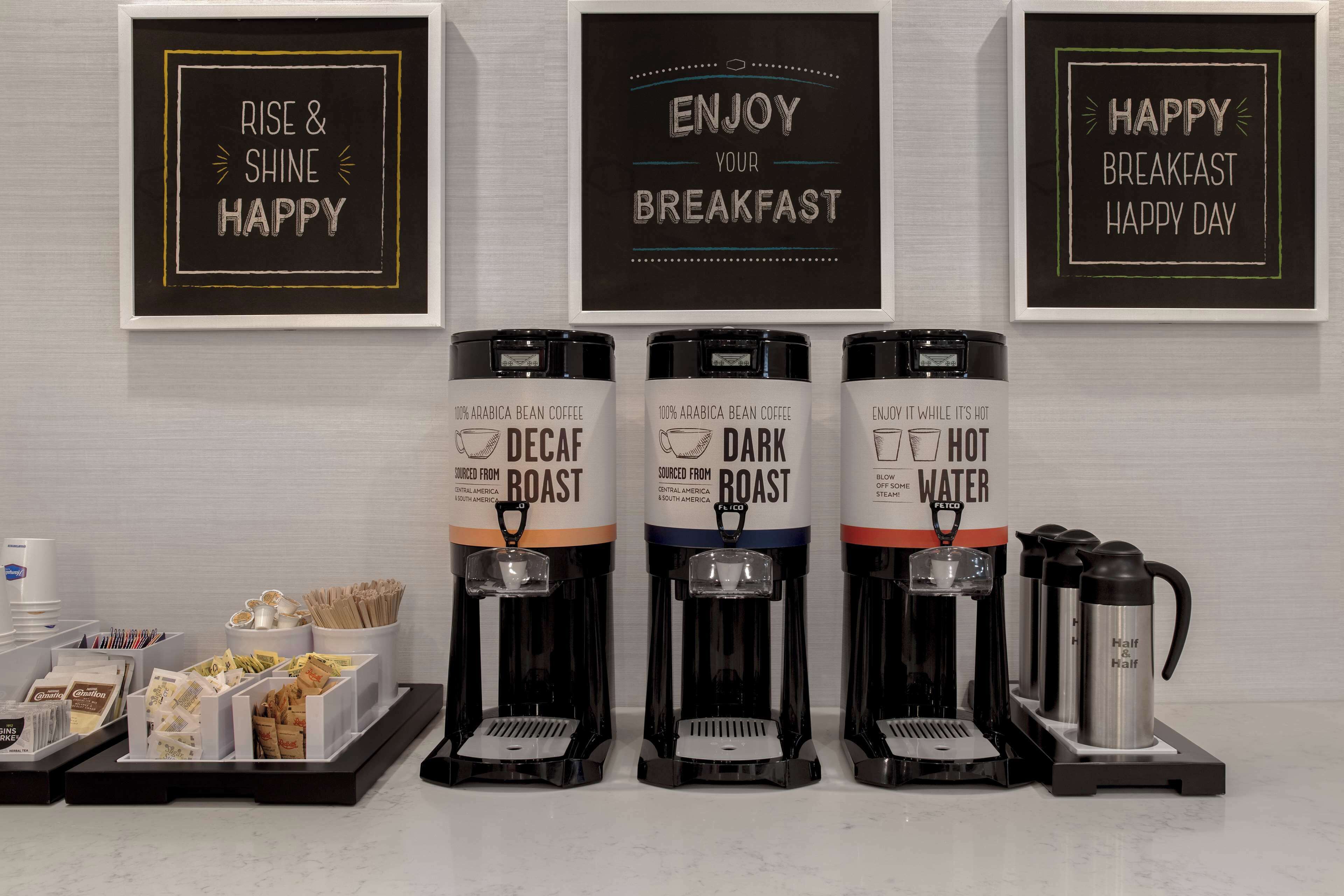 Un espace dédié au café, équipé de trois distributeurs de café étiquetés « café décaféiné », « café torréfié foncé » et « eau chaude ». On y trouve également des tasses à café et un panneau indiquant : « Bon appétit ».