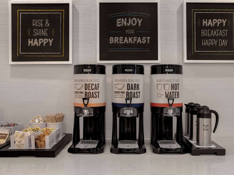 Un espace dédié au café, équipé de trois distributeurs de café étiquetés « café décaféiné », « café torréfié foncé » et « eau chaude ». On y trouve également des tasses à café et un panneau indiquant : « Bon appétit ».