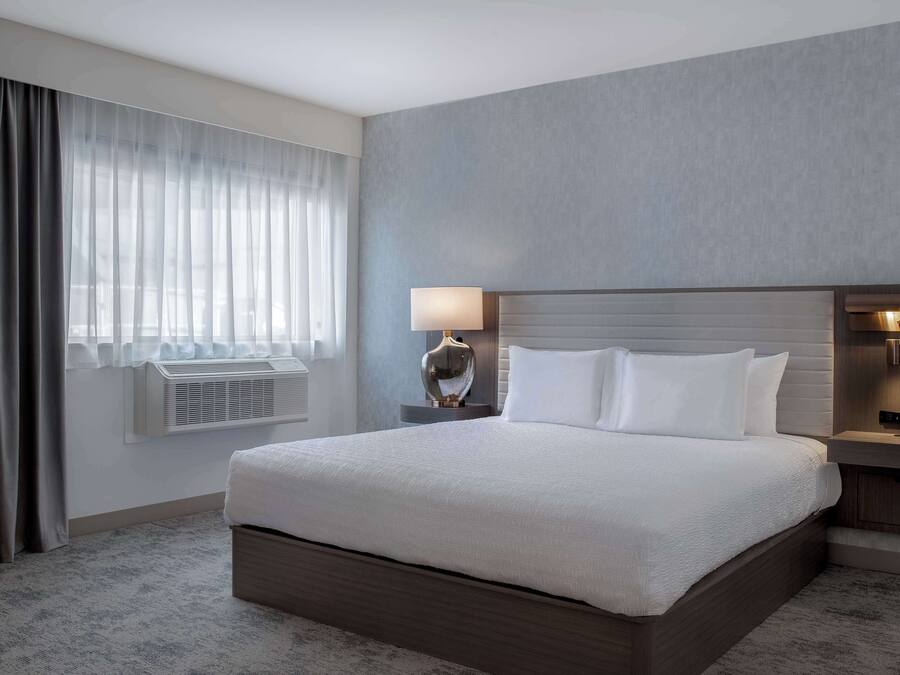 A hotel room with a large bed, a bedside table with a lamp, a wall-mounted air conditioner, and a window with curtains.