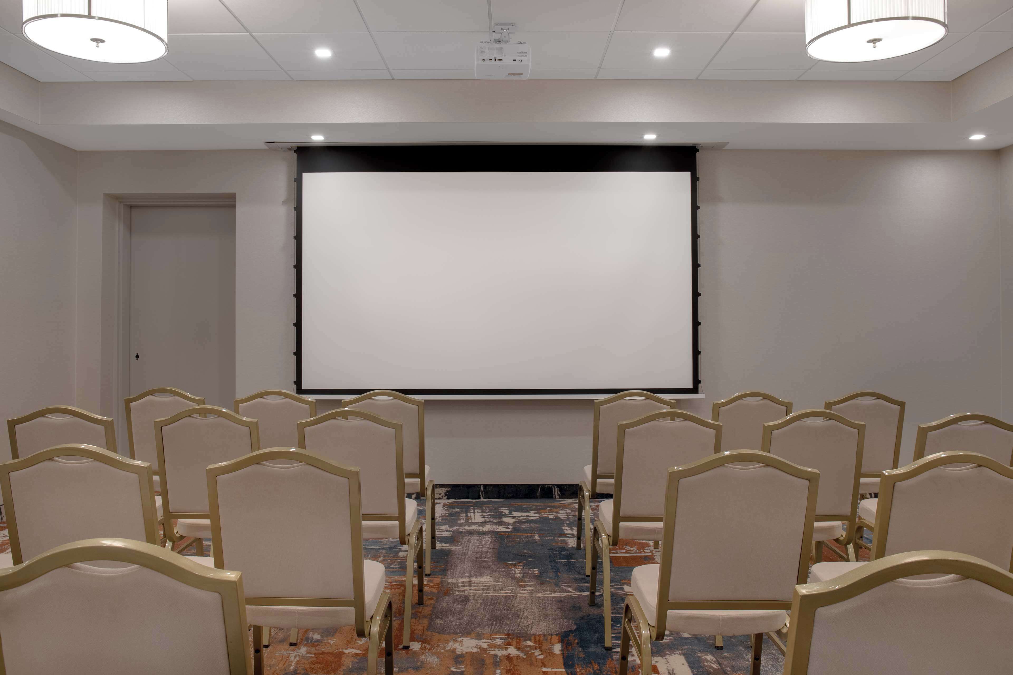 Un salón de conferencias con filas de sillas orientadas hacia una pantalla proyectora vacía, suelo alfombrado e iluminación en el techo.