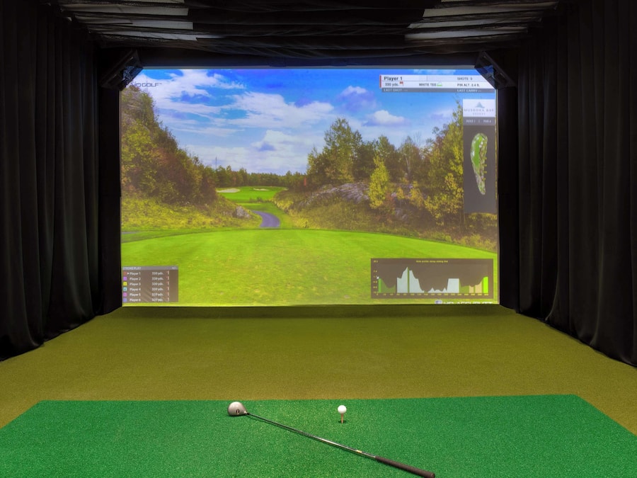 A virtual golf simulation setup with a screen displaying a golf course, a golf ball, and a club on a green mat.