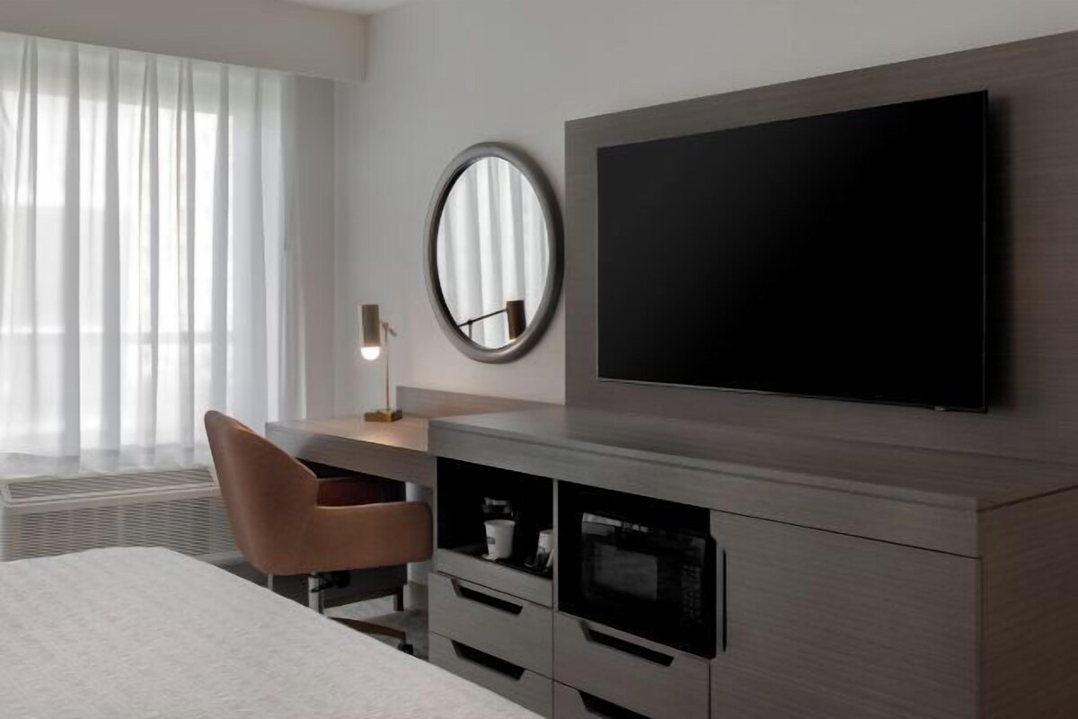 A modern hotel room with a large bed, a desk with a chair, a flat-screen TV mounted on the wall, a round mirror, and built-in cabinetry with a microwave.