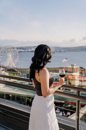 A woman holding a glass of wine, overlooking a waterfront with a Ferris wheel and a building with "PUBLIC ARCADE" signage.