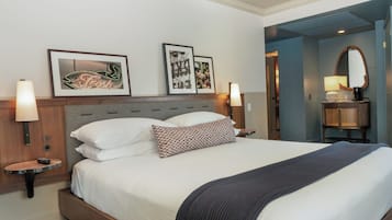 Habitación de hotel con cama, dos lámparas de noche, mesitas de noche, una pequeña mesa, un espejo y dos cuadros en la pared.