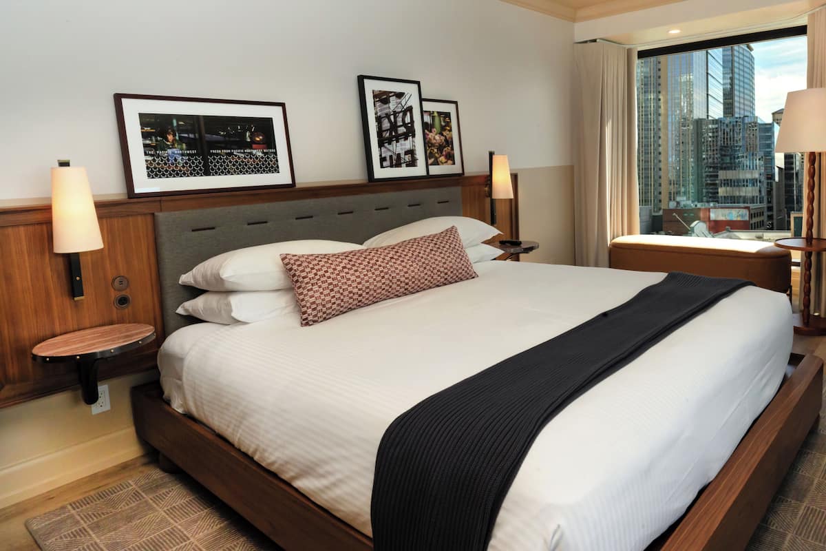 A hotel room with a large bed, two side tables, a nightstand, a window with a view of the city, and framed pictures on the wall.