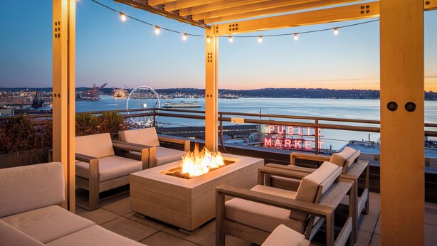 An outdoor seating area with a fire feature, overlooking a waterfront with a Ferris wheel and a sign reading "PUERTO MARKE".