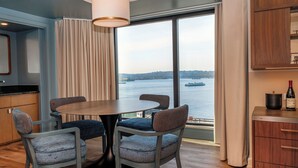 A modern room with a round table and four chairs, a large window with a view of a body of water and a boat, and a minibar with a bottle of wine.