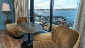 A room with a large window offering a view of a waterfront area, including a Ferris wheel and a pier. There is a round table and two chairs in the room.