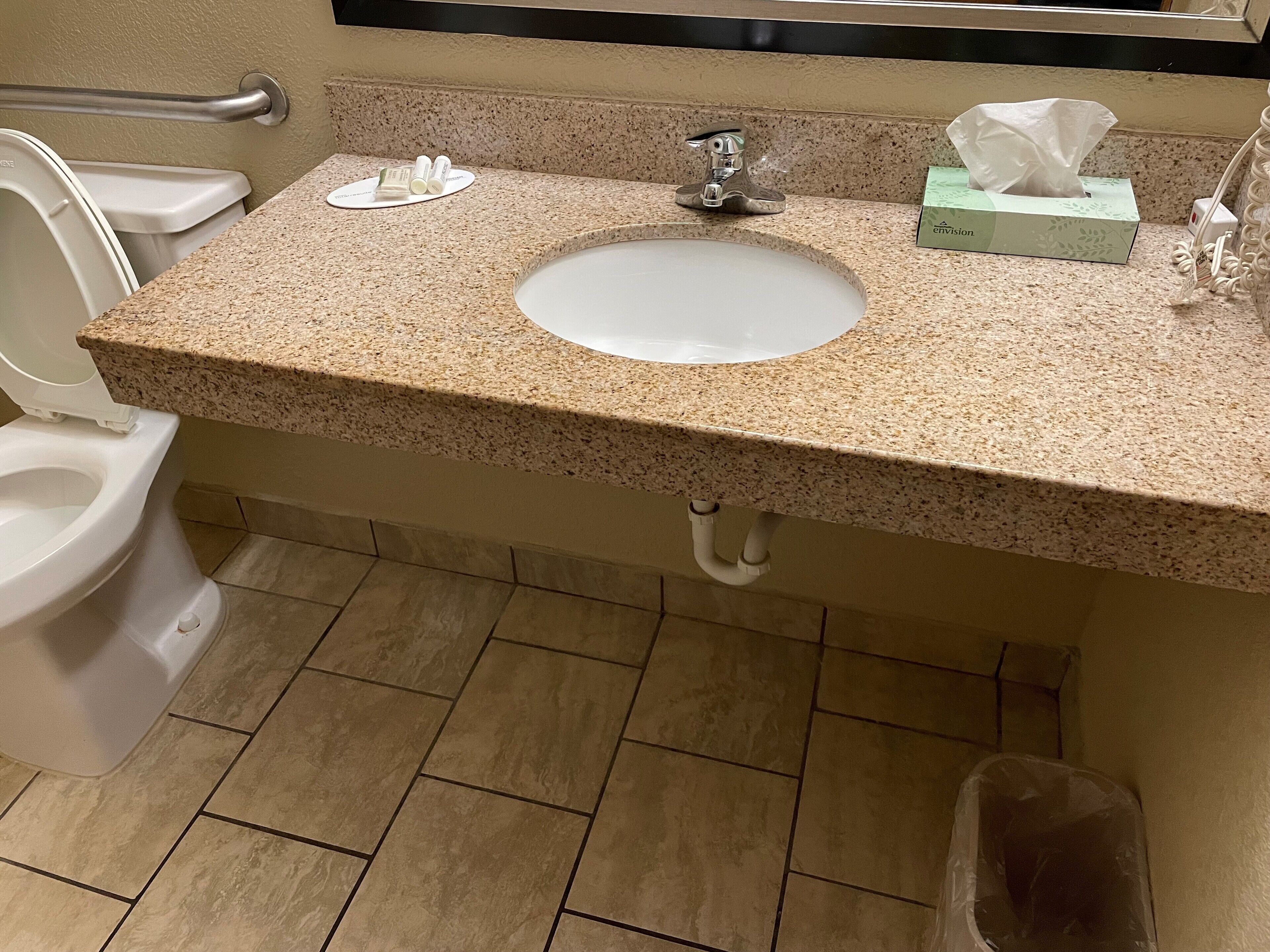 Une salle de bain avec un comptoir en granit, un lavabo blanc, un miroir, des toilettes et une poubelle.
