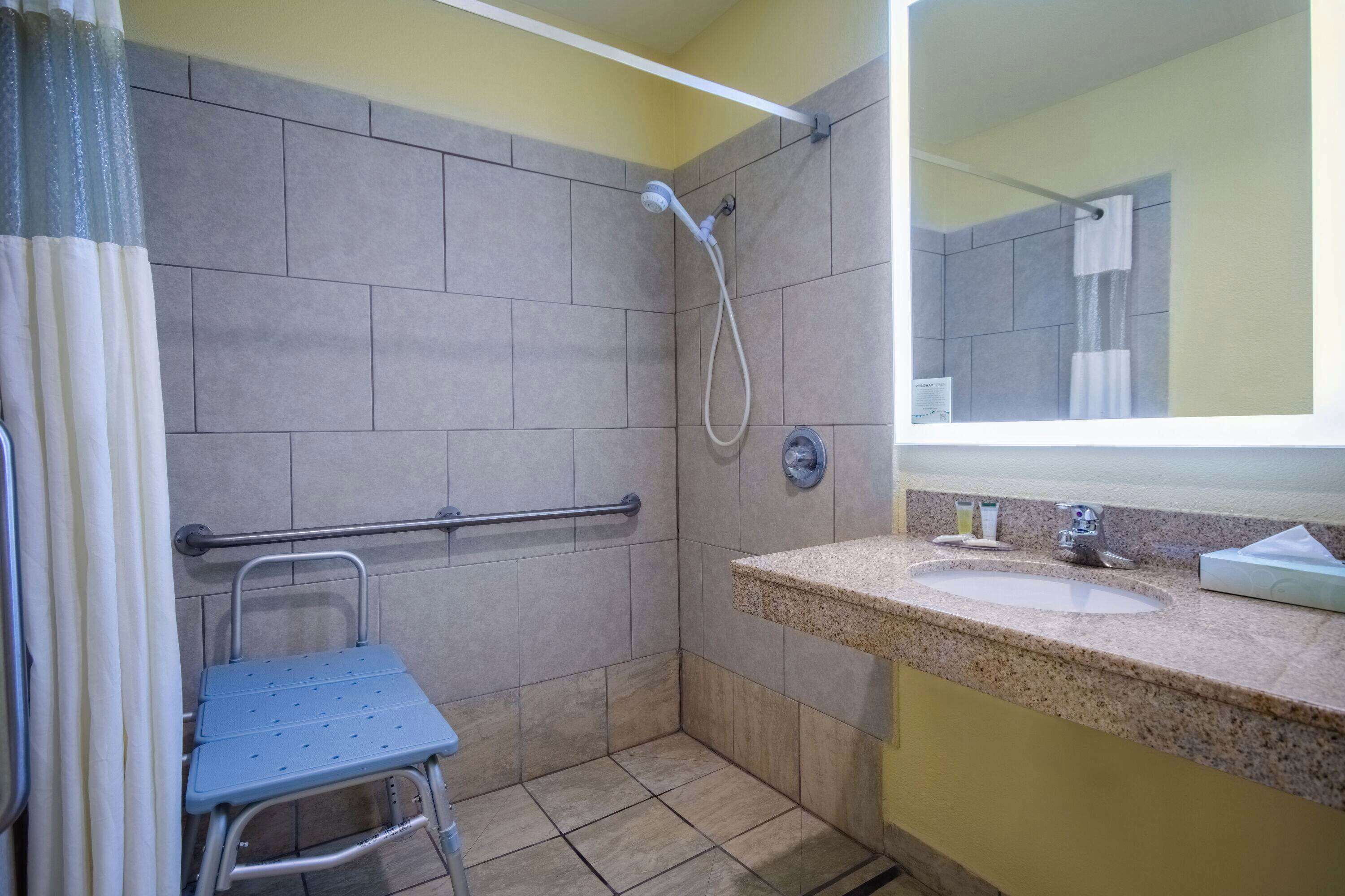 A bathroom with a shower, a sink, a mirror, and a wheelchair-accessible bench.